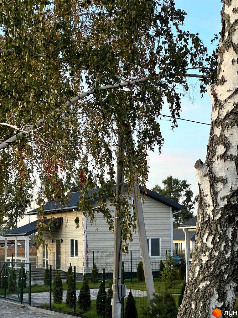 Вигляд котеджу в Містечку котеджів Lagom Village. Двоповерховий будинок зі світлим фасадом та темним дахом, зелений газон та дерева на подвір'ї.