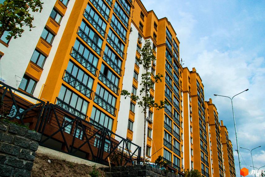 Житловий комплекс Щасливий на Петропавлівці: яскрава жовто-біла новобудова з численними вікнами та балконами, оточена деревами та огороджена парканом. На фоні блакитне небо з хмарами та ліхтарні стовпи.