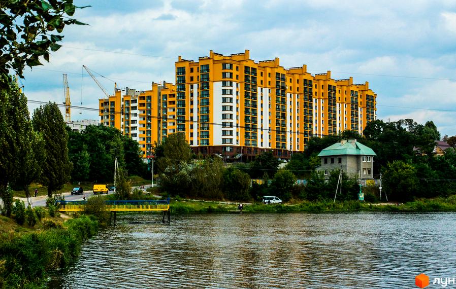Житловий комплекс Щасливий на Петропавлівці – яскрава жовто-біла новобудова біля річки. Навколо зелені дерева, дорога з автомобілями та міст.