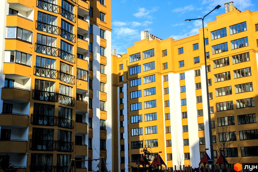 Житловий комплекс Щасливий на Петропавлівці – жовто-білі багатоповерхові будинки з балконами під блакитним небом. Поруч вуличне освітлення та елементи дитячого майданчика.