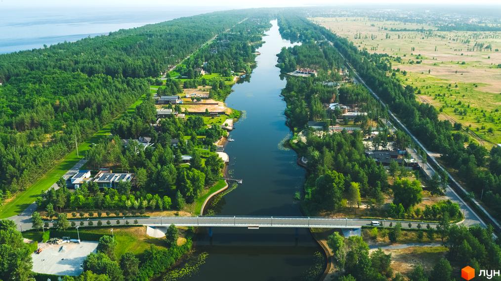 Панорамний вид на Котеджне містечко Riviera Village з річкою та мостом. Будинки серед зелених насаджень, поруч поле.