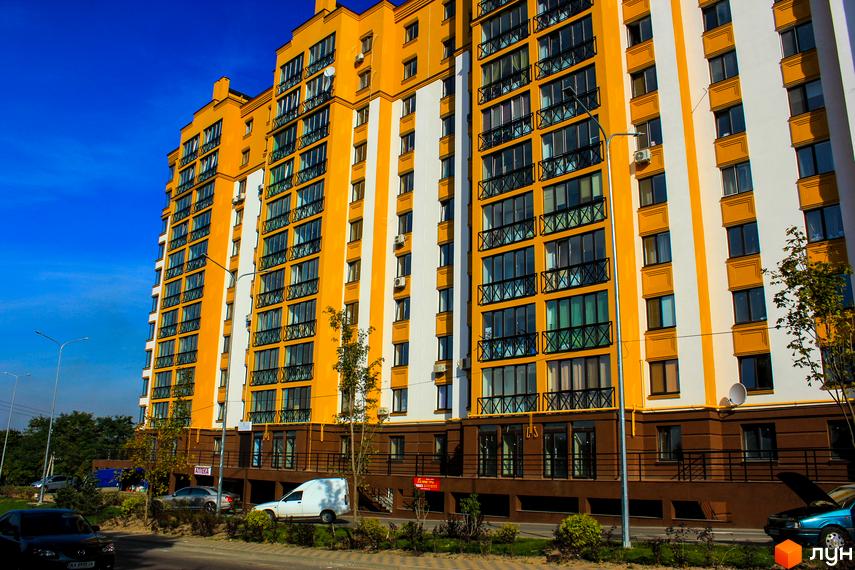 Житловий комплекс Щасливий на Петропавлівці – яскрава жовто-біла новобудова з численними вікнами та балконами. Навколо будинку розташовані зелені насадження та проїжджа частина з автомобілями.