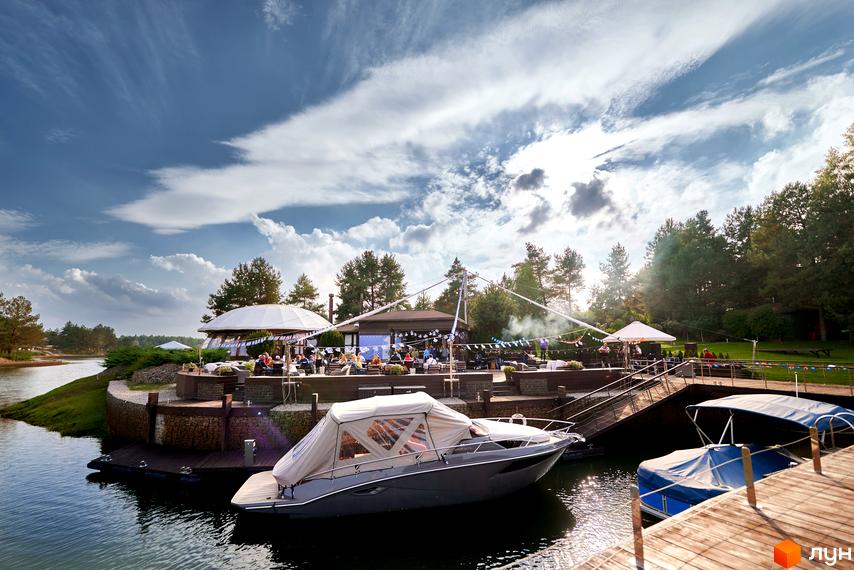 Зображення Котеджного містечка Riviera Village з терасою на березі річки. На передньому плані човен, далі - будівлі з відпочиваючими людьми.