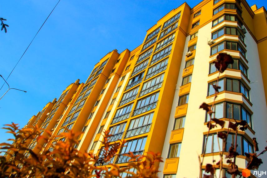 Житловий комплекс Щасливий на Петропавлівці: жовта багатоповерхівка з численними вікнами та балконами на тлі блакитного неба. На передньому плані - осіннє листя.