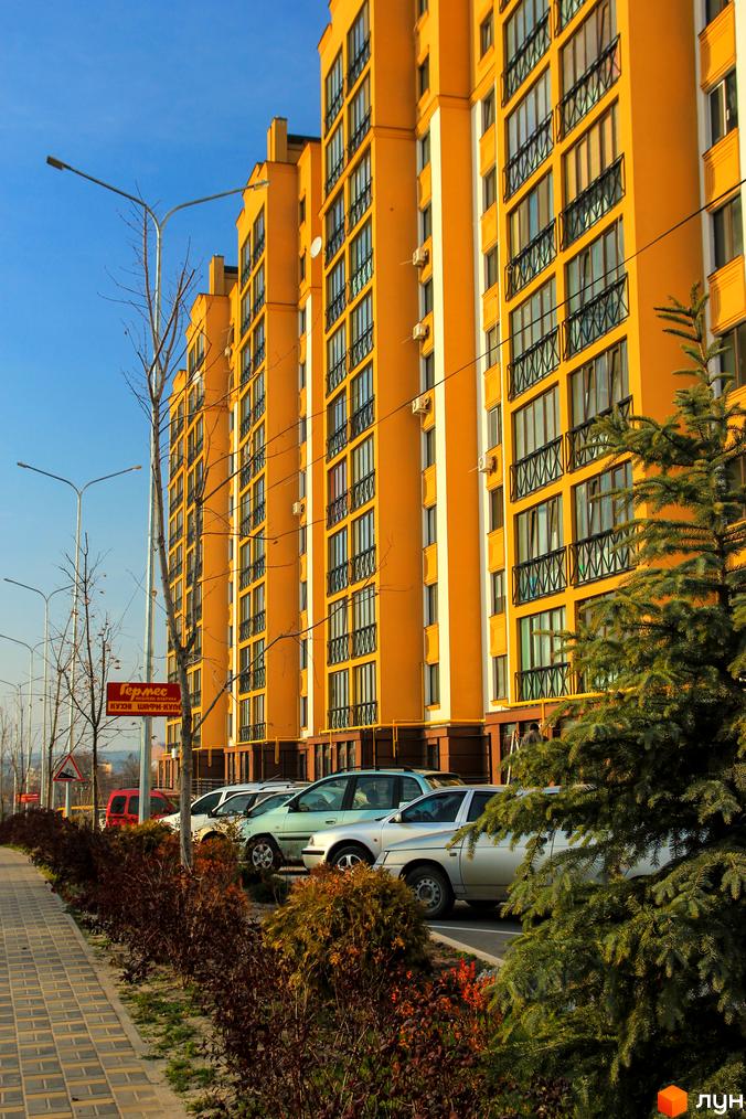 Житловий комплекс Щасливий на Петропавлівці: яскрава жовта новобудова з балконами та кондиціонерами. Поряд розташована доглянута прибудинкова територія з деревами та кущами, а також припарковані автомобілі.