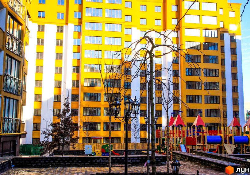 Житловий комплекс Щасливий на Петропавлівці. Яскрава жовта новобудова з білими вертикальними елементами та дитячим майданчиком.