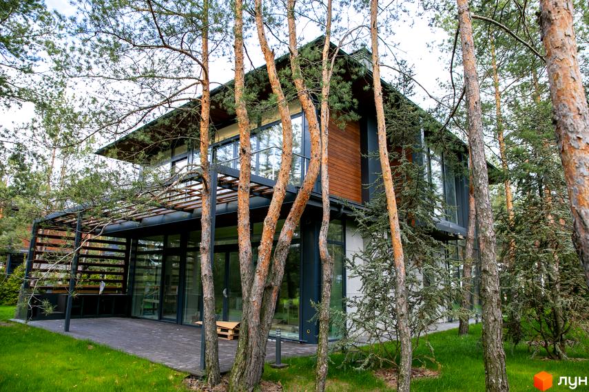Зовнішній вигляд будинку в Котеджному містечку Riviera Village. Двоповерхова будівля з великими вікнами та дерев'яним оздобленням, оточена соснами.