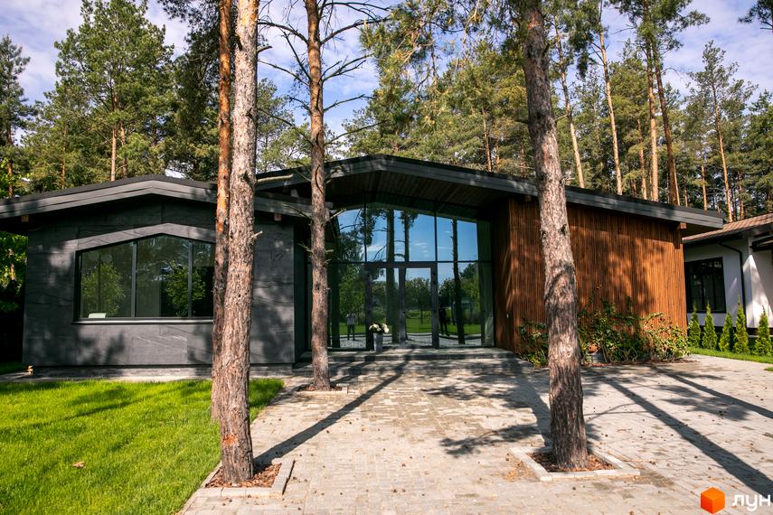 Зображення будинку в Котеджному містечку Riviera Village з великими вікнами та дерев'яною обробкою, розташованого серед сосен. На передньому плані вимощена доріжка.
