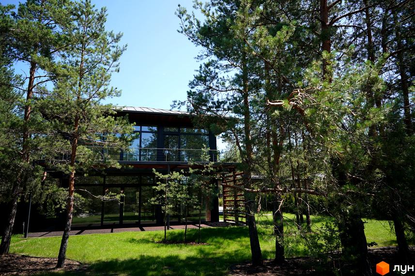 Будинок з панорамними вікнами та балконом серед дерев. На території газон та молоді дерева.