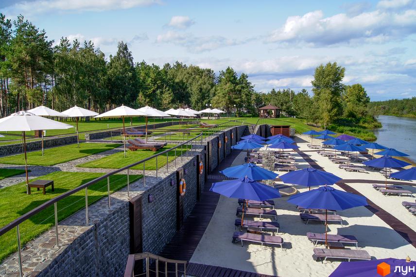 КМ Riviera Village — котеджний комплекс з білими парасольками та зоною відпочинку на березі річки. На території комплексу є зелені газони, доріжки та шезлонги.