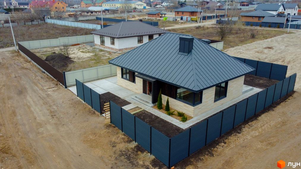 Котеджне містечко КМ SMADE Park: одноповерховий будинок з сірим дахом та цегляним фасадом, огороджений темним парканом. Навколо будинку облаштована прибудинкова територія з доріжками та зеленими насадженнями.