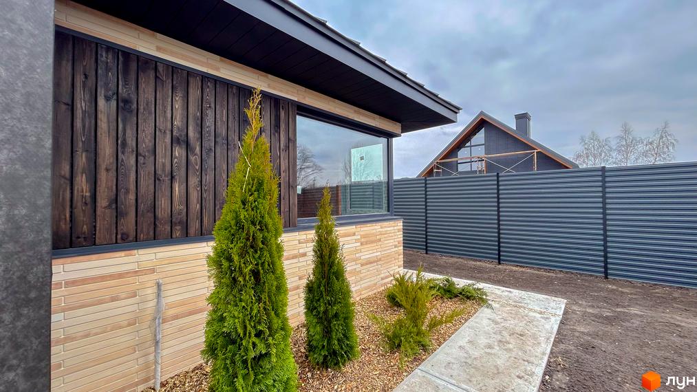 Зовнішній вигляд будинку з дерев'яним та цегляним фасадом, великим вікном та доглянутою прибудинковою територією з туями та парканом.