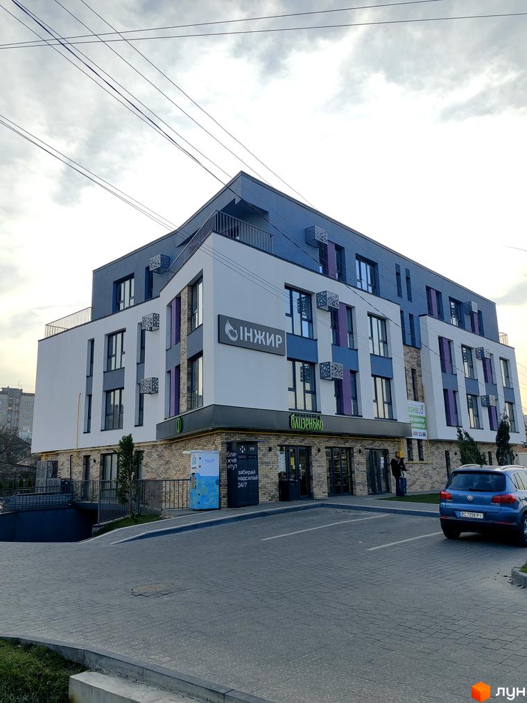 Житловий комплекс ІНЖИР - це сучасна новобудова з фасадом біло-сірого кольору та фіолетовими вставками, розташована на бруківці біля парковки.