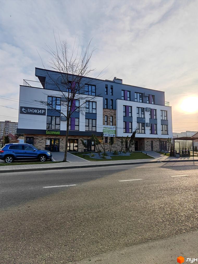 Житловий комплекс ІНЖИР - це сучасна новобудова з фасадом біло-сірого кольору, оздобленим цегляними вставками та фіолетовими акцентами. Перед будинком розташована зелена зона з деревами та доглянутим газоном.
