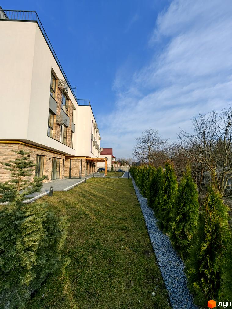 Зображення будинку в Житловому комплексі ІНЖИР з газоном та туями біля нього. На задньому плані видно інші будівлі та дерева.