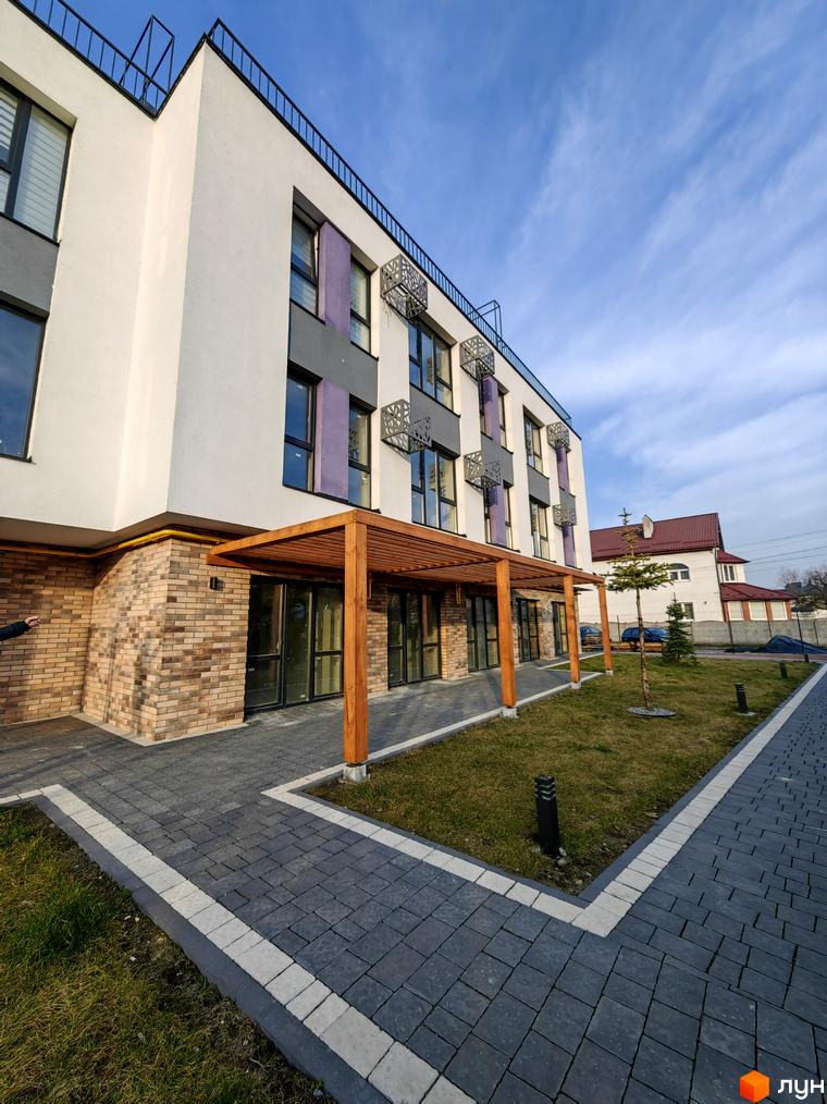 Зовнішній вигляд новобудови ЖК ІНЖИР з цегляною обробкою цоколя та дерев'яною перголою. Прибудинкова територія з бруківкою та газоном.