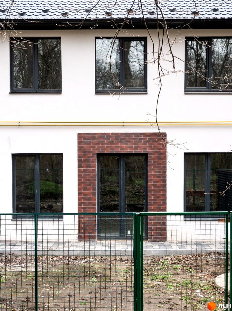 Фасад будинку з білими стінами та темними вікнами. Перед будинком зелений паркан та доріжка з плитки.