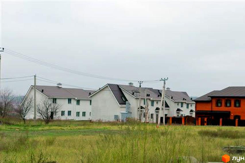 Котеджне містечко Затишний Двір: білі будинки з темними дахами та один помаранчевий будинок. Навколо зелена трава та стовпи з дротами.