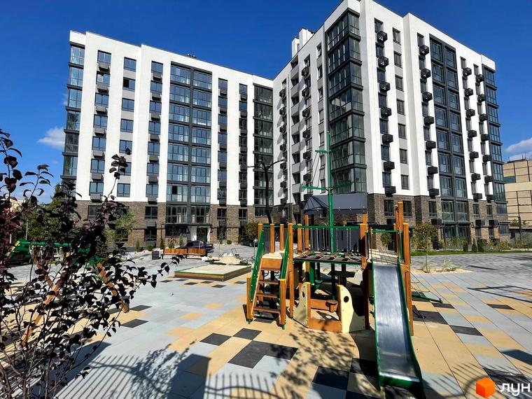 Житловий комплекс Sweet Home з білою фасадною частиною та великою кількістю вікон. На прибудинковій території розташований дитячий майданчик з гіркою та пісочницею.