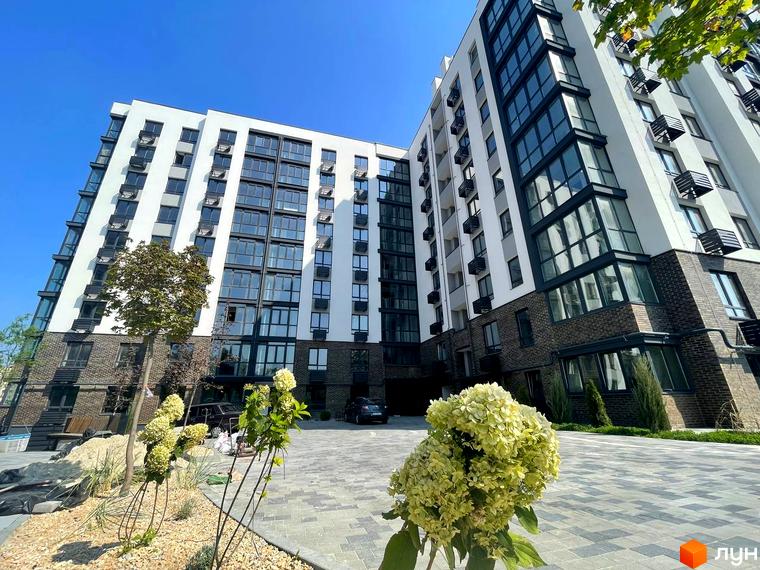 Житловий комплекс Sweet Home з багатоповерховим будинком, що має білий фасад, темну цеглу та похилі архітектурні елементи. Прибудинкова територія облаштована бруківкою та зеленими насадженнями.