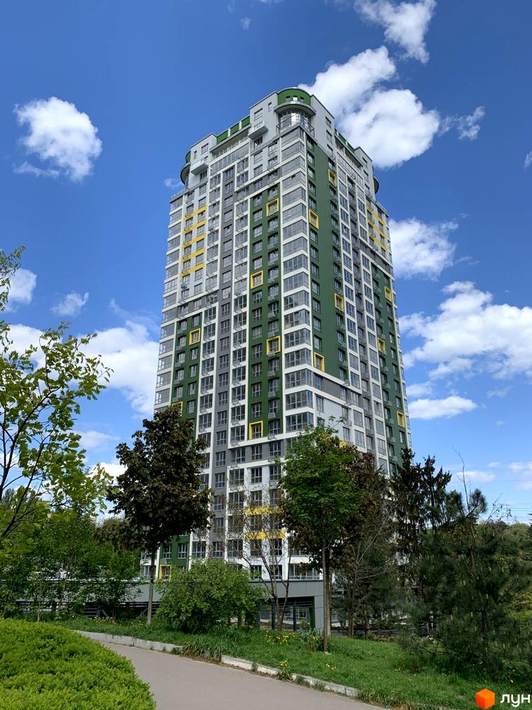 Житловий комплекс Паркове місто: високий будинок з фасадом зеленого, сірого та жовтого кольорів. Прибудинкова територія озеленена деревами та кущами, є доріжка.