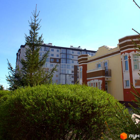 Житловий комплекс Idilika Avenue: багатоповерхова новобудова з панорамними вікнами та сусідня будівля з жовто-коричневим фасадом на фоні блакитного неба. Попереду зелені кущі та дерева.