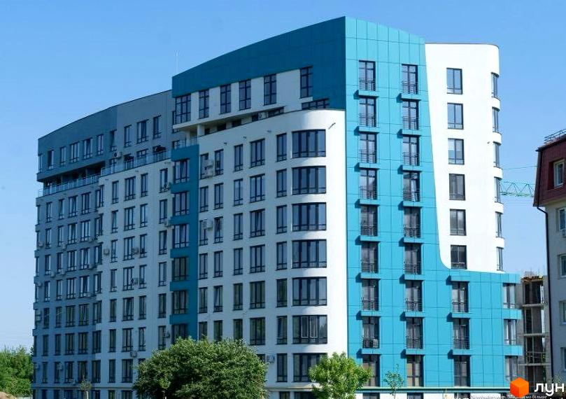 Зовнішній вигляд житлового комплексу Парус Riverside з біло-блакитними фасадами та великою кількістю вікон. Навколо новобудови зелені насадження та блакитне небо.