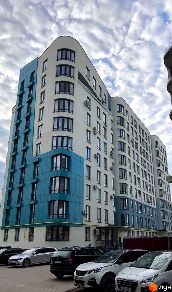 Житловий комплекс Парус Riverside з білою та блакитною фасадною обробкою та великою кількістю вікон. Перед будівлею припарковані автомобілі.