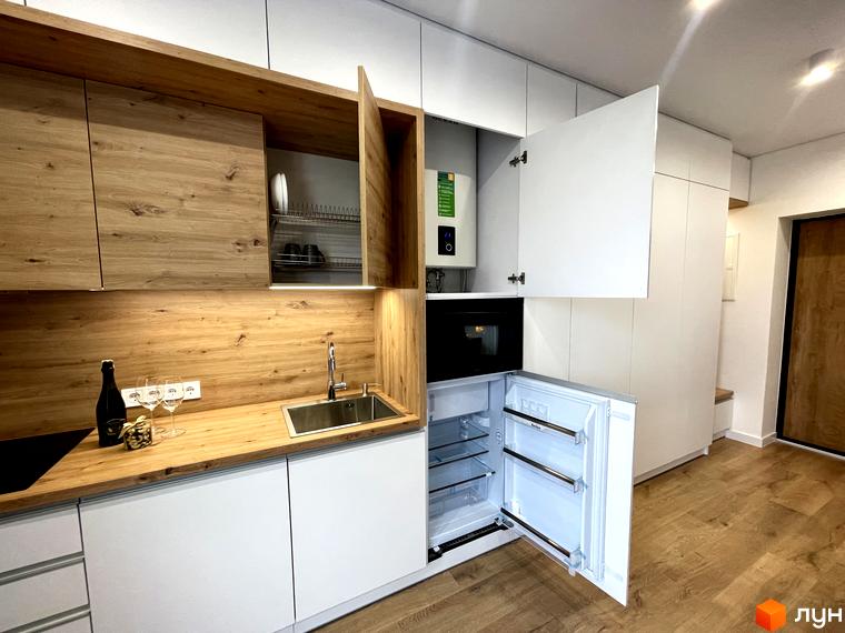 Функціональна кухня-студія в апартаментах Coliving EST з дерев'яними фасадами, вбудованою побутовою технікою та компактним холодильником.