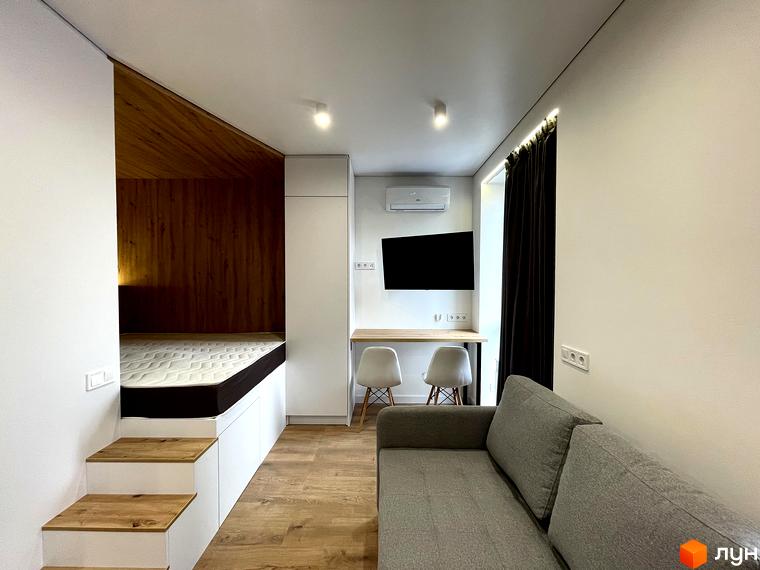 Інтер'єр студії з окремою спальною зоною, облаштованою двоспальним ліжком та дерев'яними елементами. Поруч розташована робоча зона з письмовим столом та двома стільцями.