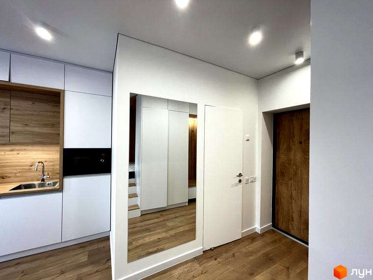 Інтер'єр квартири в Coliving EST: кухня з дерев'яними елементами та білими шафами, передпокій з дзеркалом та дерев'яними вхідними дверима.