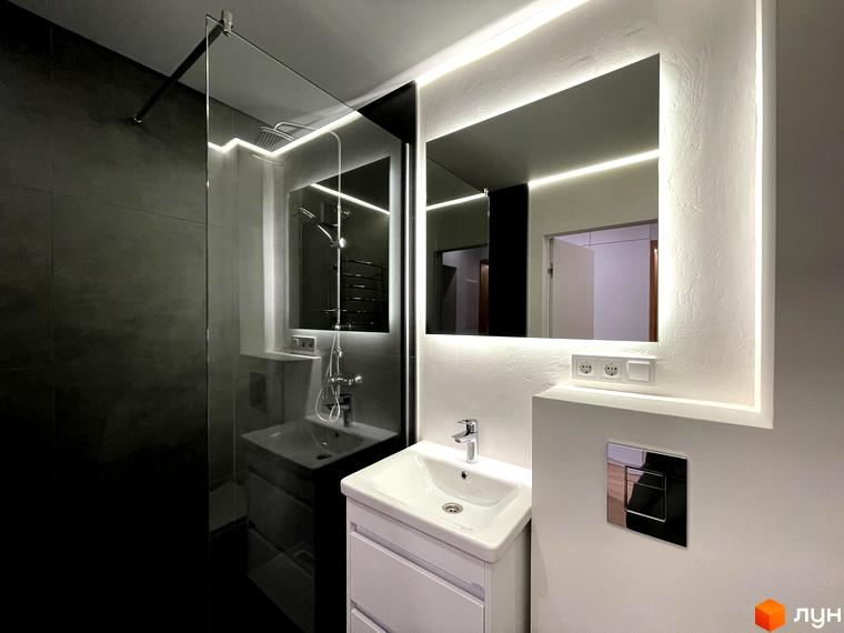 Інтер'єр ванної кімнати в Coliving EST: душова зона з темною плиткою, дзеркало з підсвічуванням, біла тумба з раковиною.