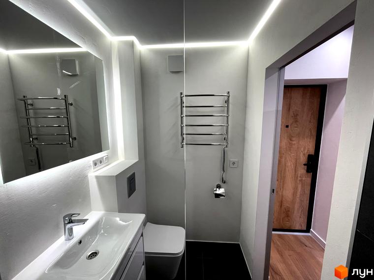 Інтер'єр санвузла в Coliving EST з підсвіченим дзеркалом, раковиною, унітазом та рушникосушкою. Видно дерев'яні двері до іншої кімнати.