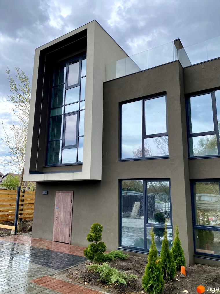 Будинок Elwood Townhouse з великими вікнами та скляним огородженням тераси. Навколо будинку облаштована прибудинкова територія з бруківкою та туями.