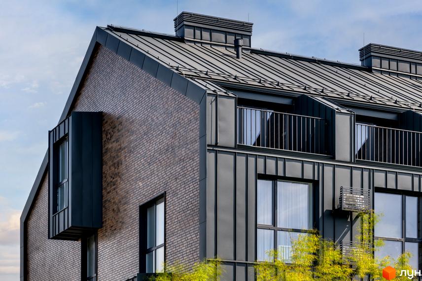 Фасад житлового комплексу Coliving Akadem з темно-сірою металевою покрівлею та комбінованим оздобленням з цегли і металевих панелей, оточений зеленою рослинністю.