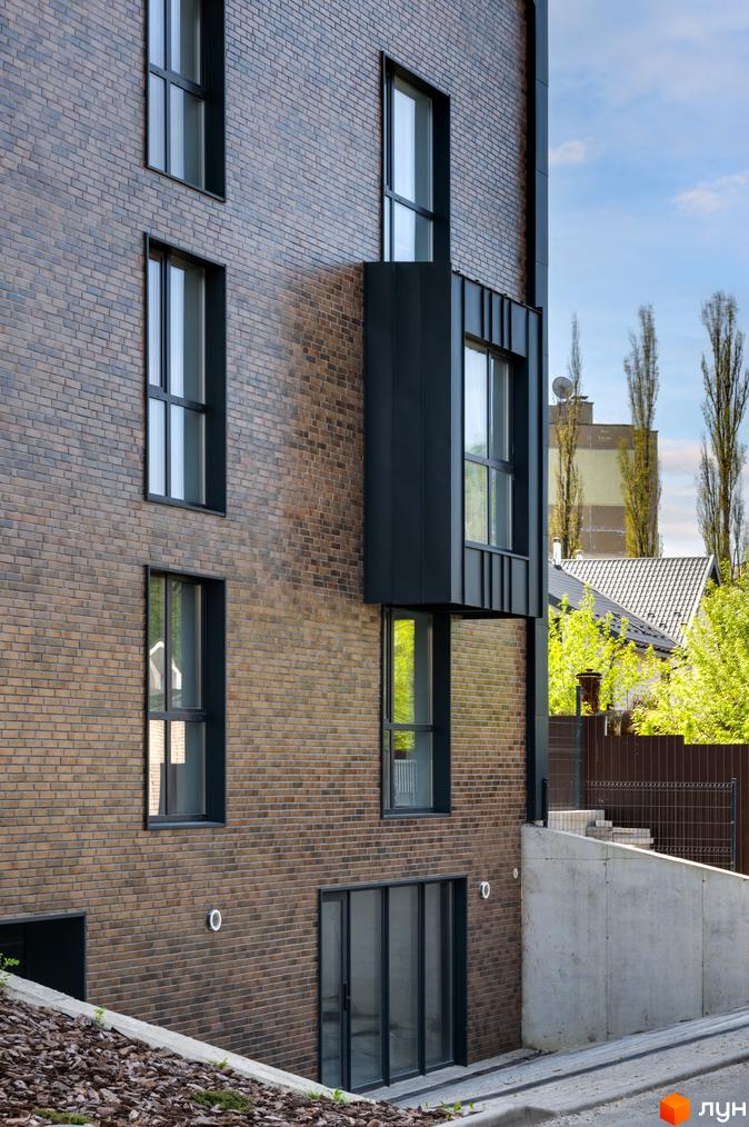 Фасад житлового комплексу Coliving Akadem з цегли, темними вікнами та виступаючим елементом. Прилегла територія має бетонну стіну та зелені насадження.