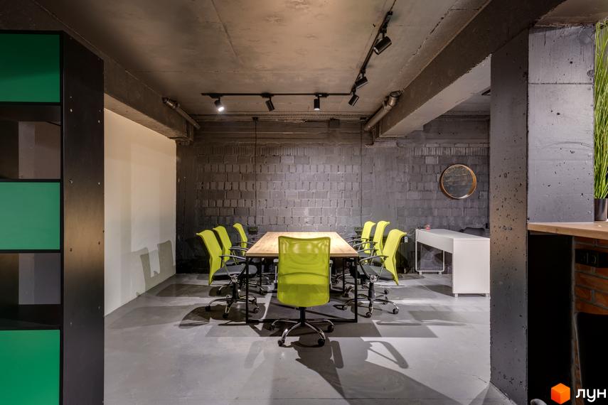 Переговорна кімната в Coliving Akadem з дерев'яним столом та салатовими кріслами. Приміщення має сірі цегляні стіни та бетонну стелю.