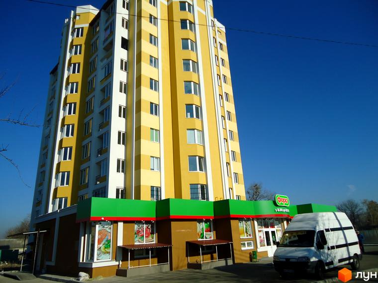 Житловий комплекс Гостомельський квартал з жовто-білим фасадом та зеленим цоколем. Поруч розташований магазин та білий фургон.