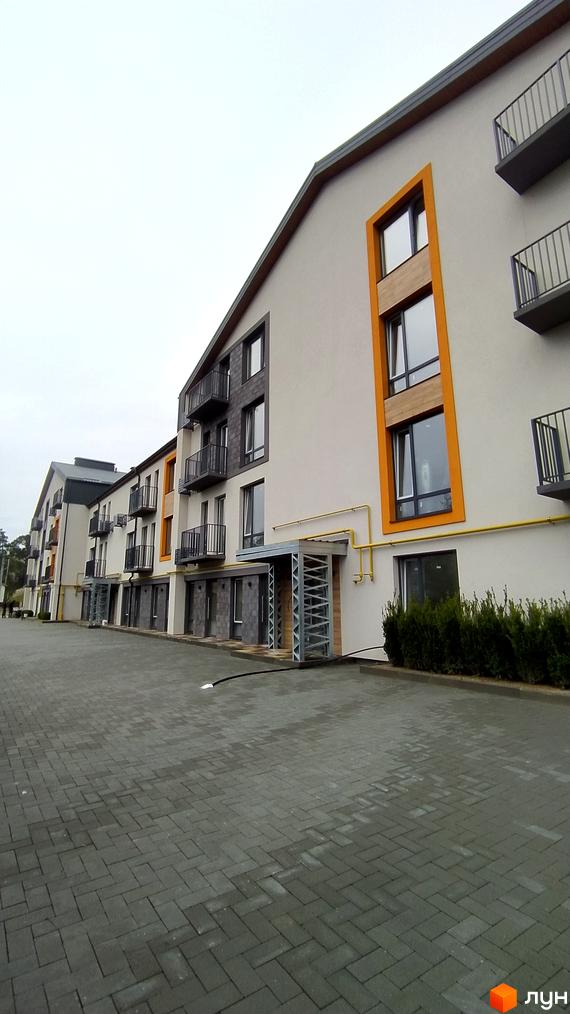 Новобудова на вул. Києво-Мироцька, 99а з сірим фасадом та оранжевими елементами. Перед будинком вимощена сіра бруківка та зелені кущі.