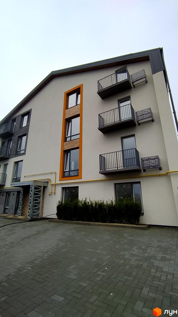 Фасад житлового комплексу по вул. Києво-Мироцька, 99а, з балконами та вікнами, на тлі сірого неба. Перед будинком розташована прибудинкова територія з бруківкою та кущами.