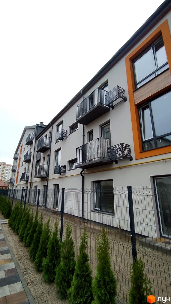 Зовнішній вигляд фасаду житлового комплексу на вул. Києво-Мироцька, 99а, з балконами та вікнами, а також прибудинкова територія з огорожею та туями.