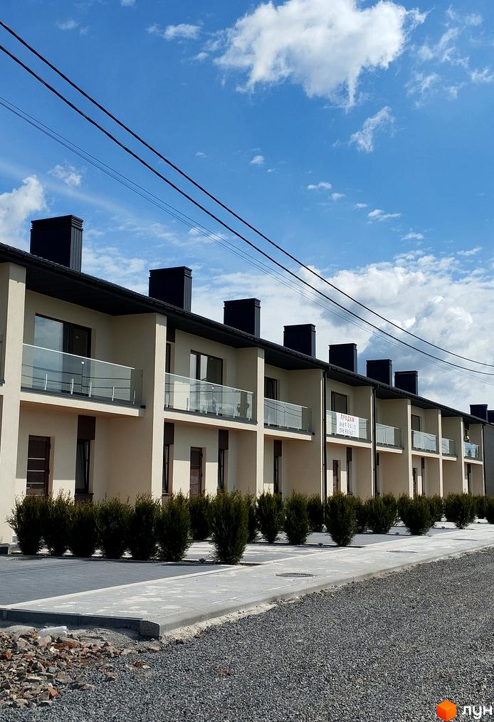 Зовнішній вигляд новобудови з балконами та димарями на фоні блакитного неба з хмарами. Прибудинкова територія з акуратно висадженими кущами та доріжками.