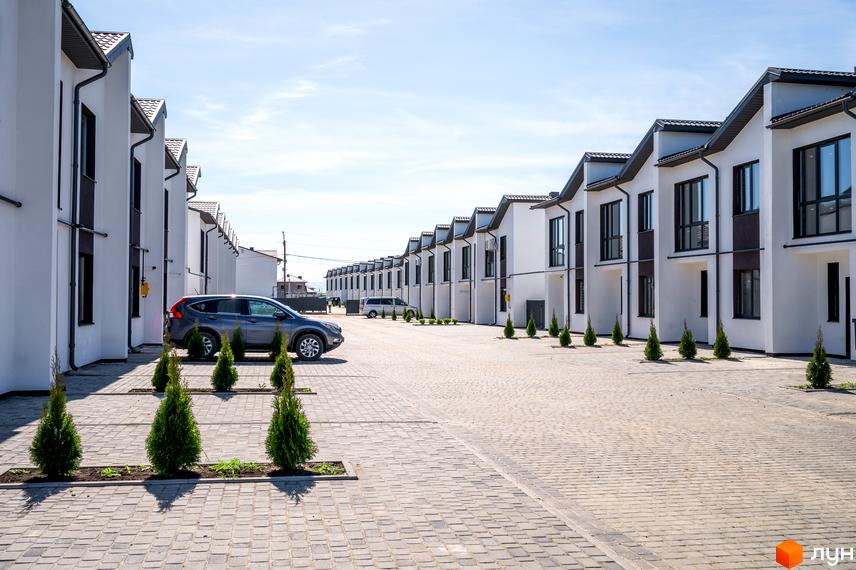 Котеджне містечко WEST TOWN: білі двоповерхові таунхауси з темними дахами та вікнами, розташовані вздовж вимощеної бруківкою дороги з зеленими насадженнями.