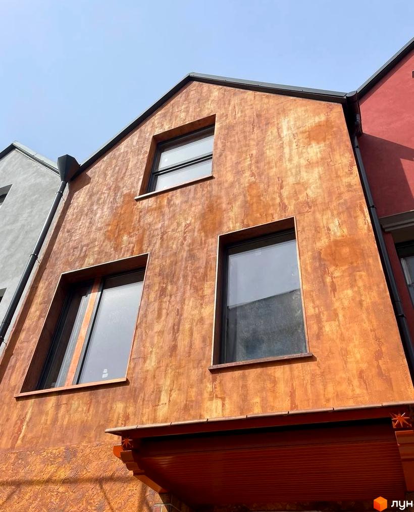 Фасад будинку в КМ КАDЕНЮК homes з вікнами та текстурною обробкою, що нагадує іржу. На сусідніх будівлях видно сіру та червону штукатурку.