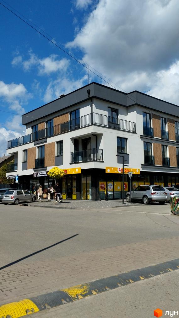 Зовнішній вигляд новобудови на вул. Олексія Бестужева-Рюміна, 7, з фасадом біло-сірого кольору та дерев'яними вставками. Перед будинком розташована прибудинкова територія з автомобілями та пішохідною зоною.