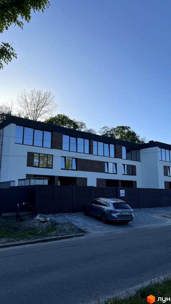 Будинок з цегляними та білими фасадами, великими вікнами, на тлі блакитного неба. Прибудинкова територія з парканом та автомобілем.