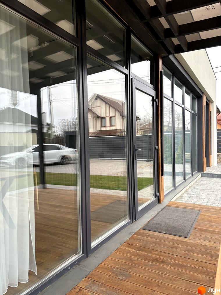 Зовнішній вигляд будинку КМ Romankiv House з великими вікнами та дерев'яною терасою. Поруч розташована прибудинкова територія з газоном та автомобілем.