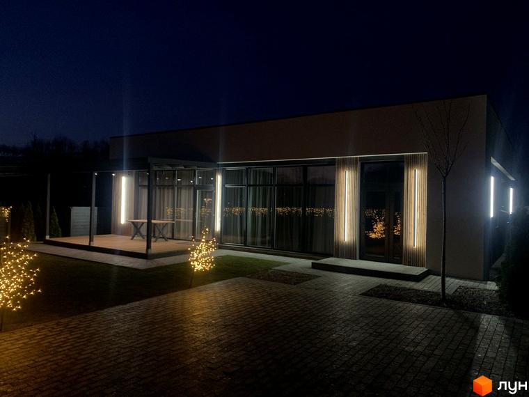 Котеджне містечко Romankiv House з підсвіченим фасадом та терасою вночі. Навколо будинку розташовані декоративні дерева з гірляндами та бруківка.