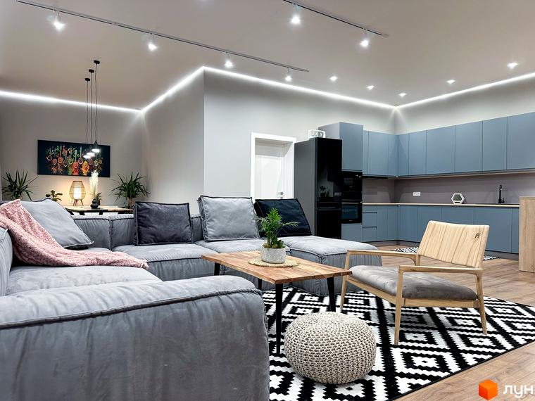 Інтер'єр вітальні з сірим диваном, дерев'яним журнальним столиком та кріслом. На задньому плані кухня з синіми шафами.