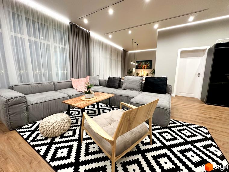 Вітальня в КМ Romankiv House з сірим кутовим диваном, дерев'яним журнальним столиком та килимом з геометричним візерунком. Простора кімната з великими вікнами та сучасним освітленням.